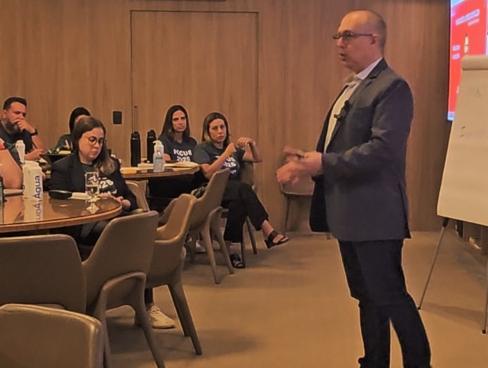 Rogério Chelucci conduzindo treinamento em sala de reunião com participantes em mesas redondas